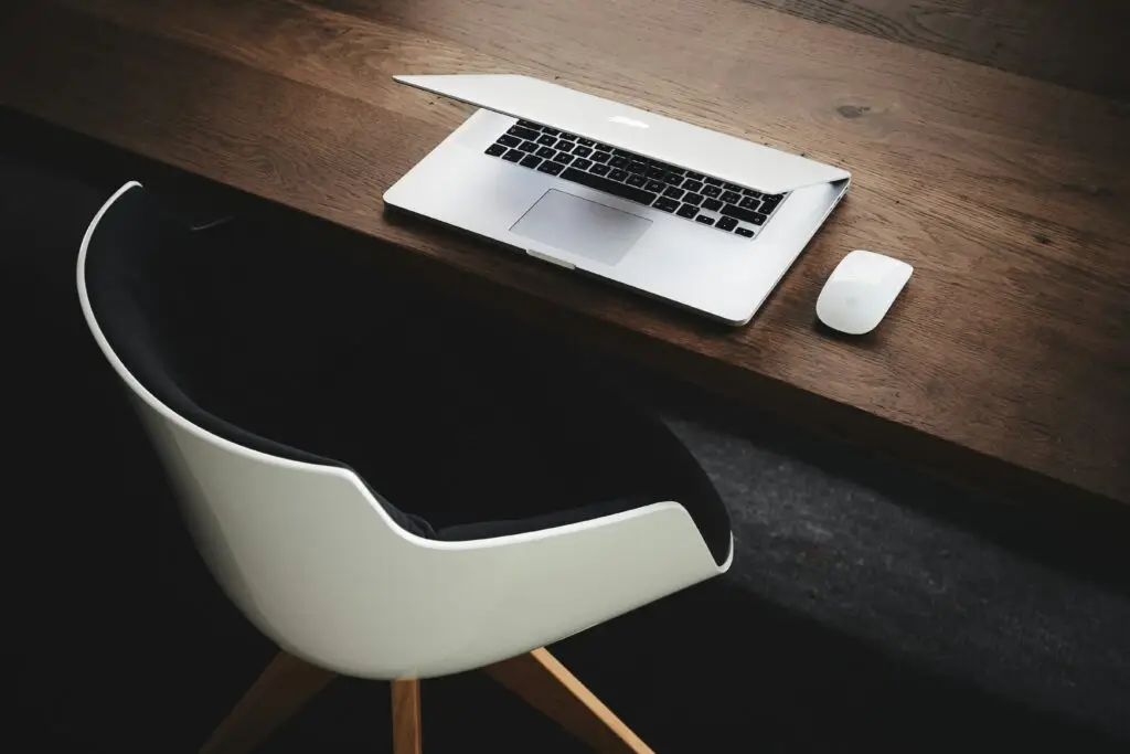 Designerstuhl vor Arbeitstisch aus Holz auf dem halb offener iMac mit Maus minimalistisch steht bereit für administrative Massnahmen Foto von Luca Bravo auf Unsplash