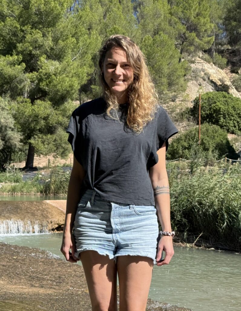 Francesca Schori in kurzen Jeans und mit schwarzem T-shirt im Wasser laufend, im Hintergrund grüne Büsche und Bäume