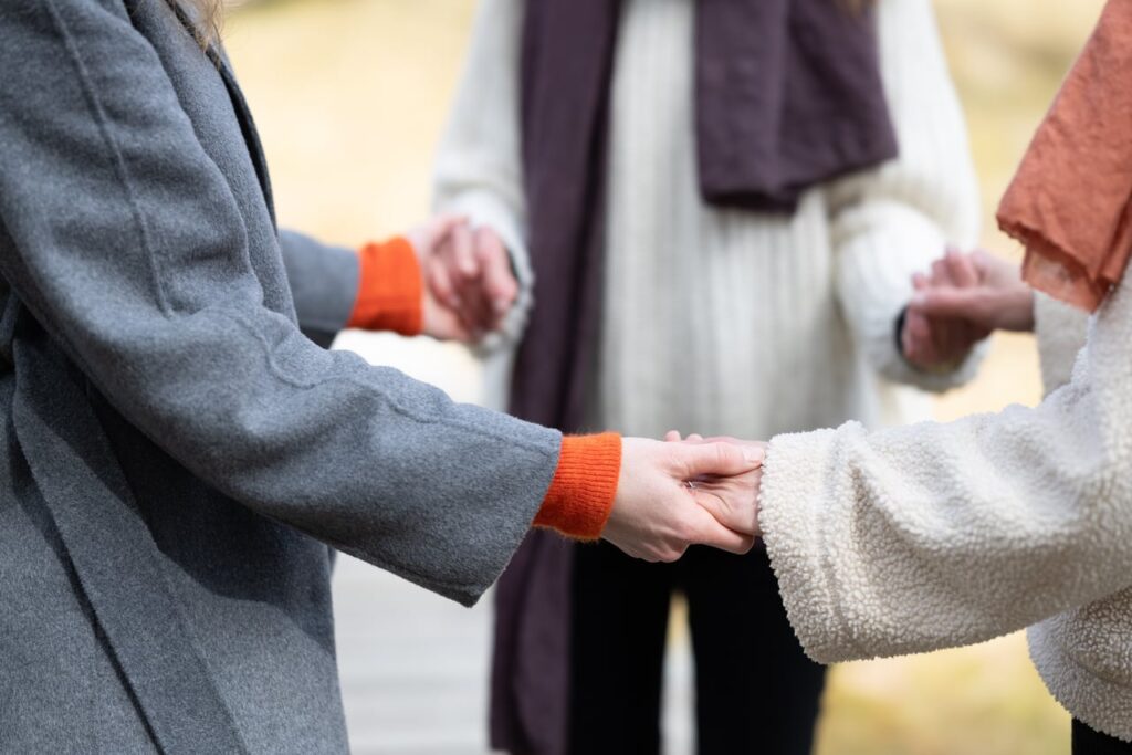 drei Frauen von denen man nur die Oberkörper sieht, stehen im Kreis und halten sich an den Händen